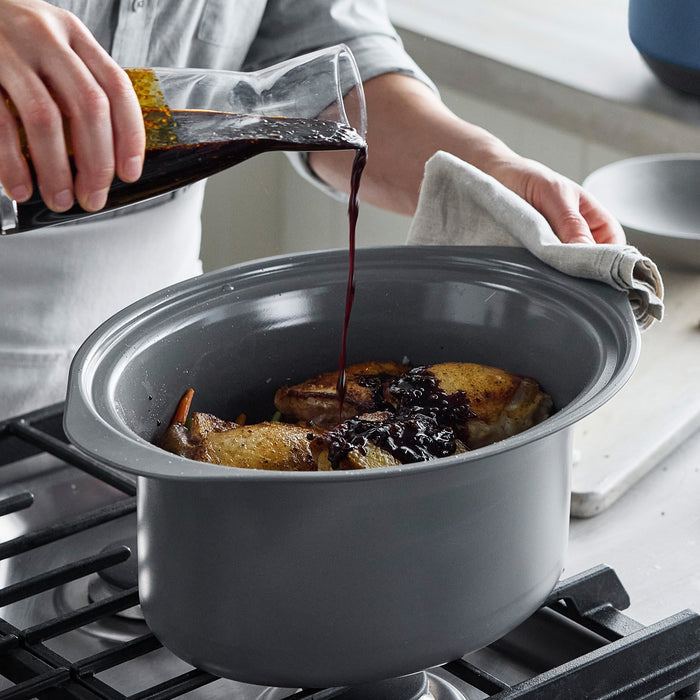 Person pouring sauce into a pot on a stove with food inside