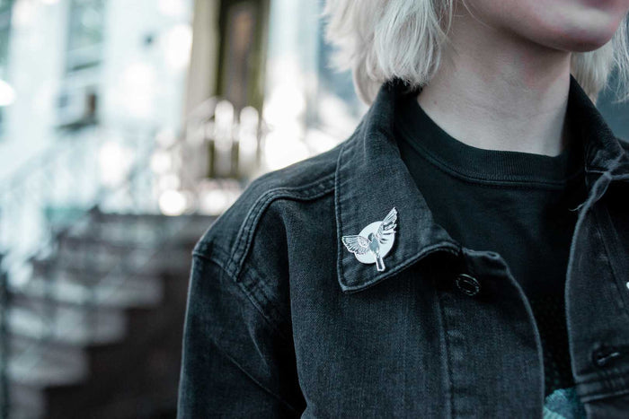 A person wears a black denim jacket by Sleep On It, featuring a bird-shaped enamel pin on the collar; blurred background shows stairs and glass structures.