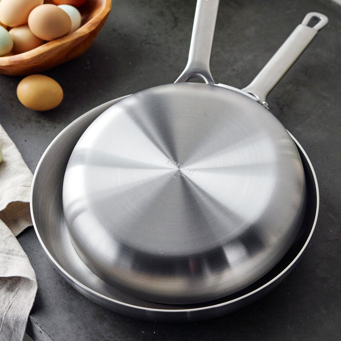Two black GreenPan Bobby Flay frypans, 10 and 12, stacked on a dark countertop with eggs in the background.
