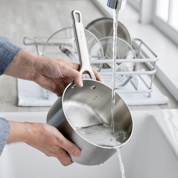 A person rinsing a stainless steel saucepan in a kitchen sink with clean dishes on the counter nearby.