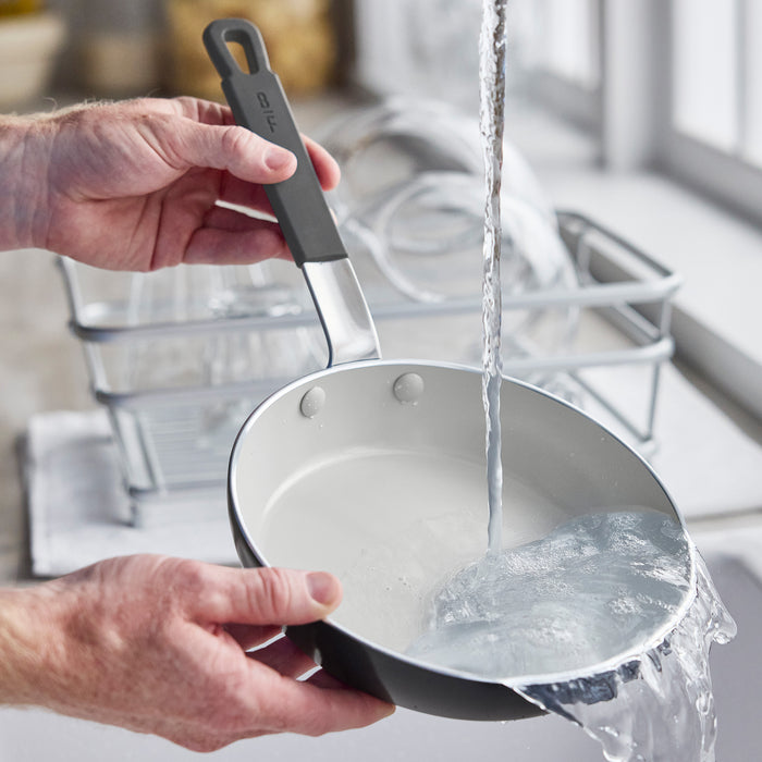 A person rinses a silver GreenPan ceramic nonstick frypan under running water near a dish rack.
