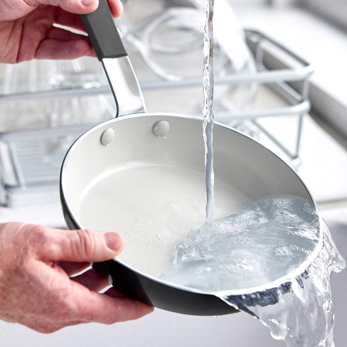 A hand rinses a silver GreenPan ceramic nonstick frypan under running water in a kitchen sink.