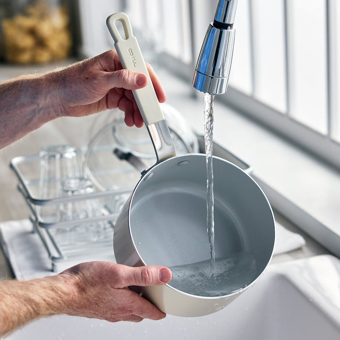 Person pouring water into a white ceramic nonstick saucepan with metal handle on a kitchen counter.