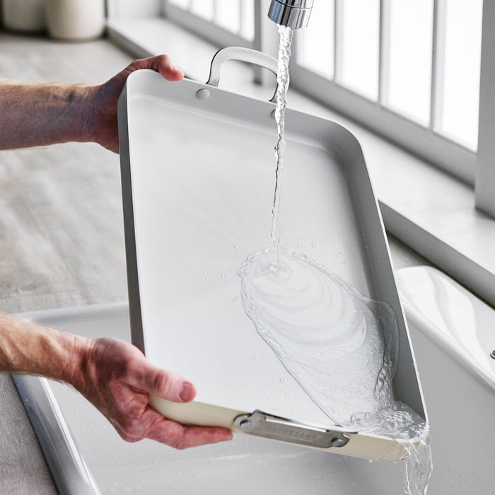 Ceramic nonstick griddle being rinsed under running water in a kitchen sink.