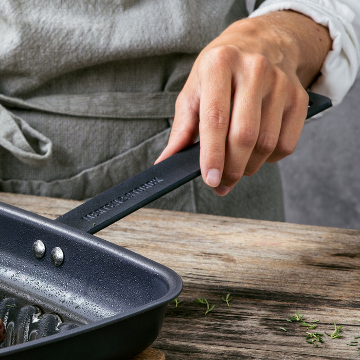 Person in an apron holding an 11-inch carbon steel square grill pan on a rustic wooden table.