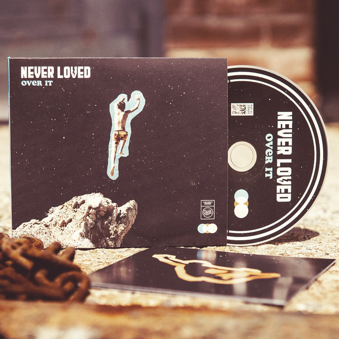 Square CD with NEVER LOVED written in the top left corner in white font. Below that there is blue text that says OVER IT. The album artwork of a boy jumping off of a cliff, but in a starry space sky. There is a black disc peeking out of the side with the same text on it, all written in white. Both are standing up on a concrete surface with rusted chains in the corner.