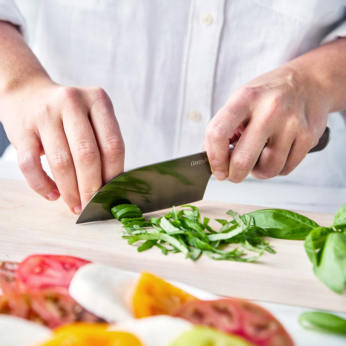 Person slices basil on a cutting board with a Premiere Titanium Cutlery 5-inch small Santoku knife, wearing a white shirt.