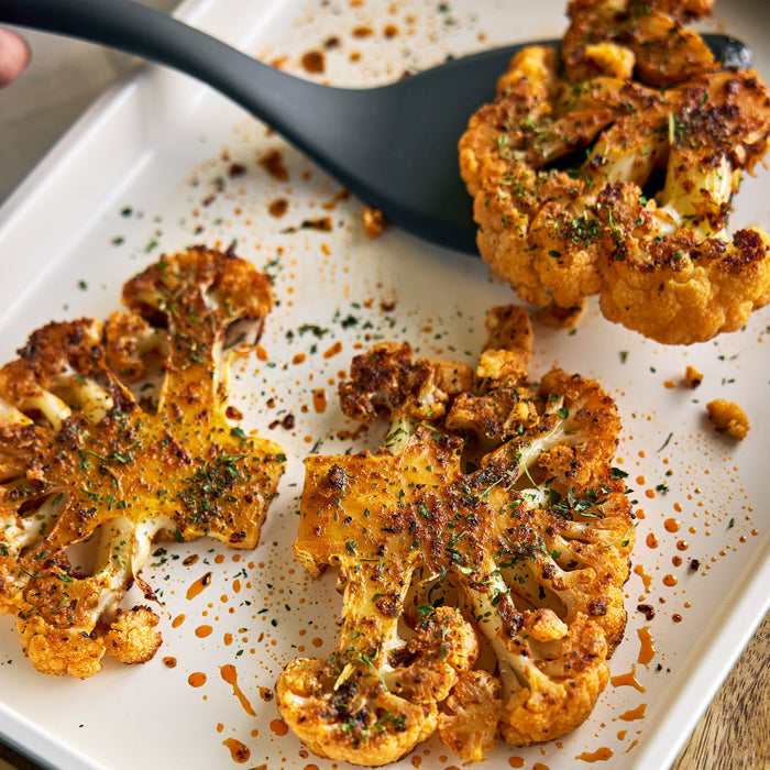 Three cauliflower steaks on a dark Reserve Bakeware tray from the 8-piece set, one lifted by a black spatula.