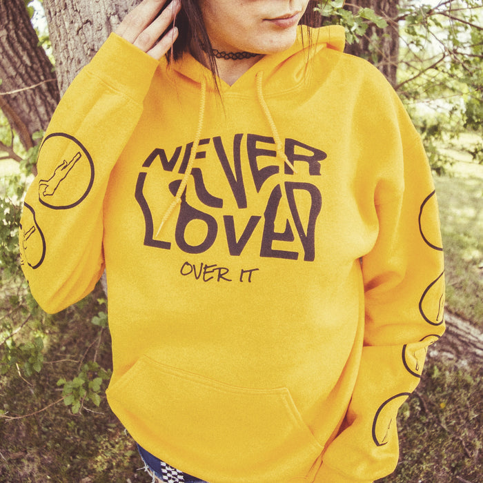 Gold colored pullover hoodie with NEVER LOVED written across the chest in wavy black font. Below that there is smaller black text that says OVER IT. On both sleeves there are cutouts of a person jumping in the air, surrounded by a circle. An individual is modeling the hoodie and standing in front of a tree in a forest.