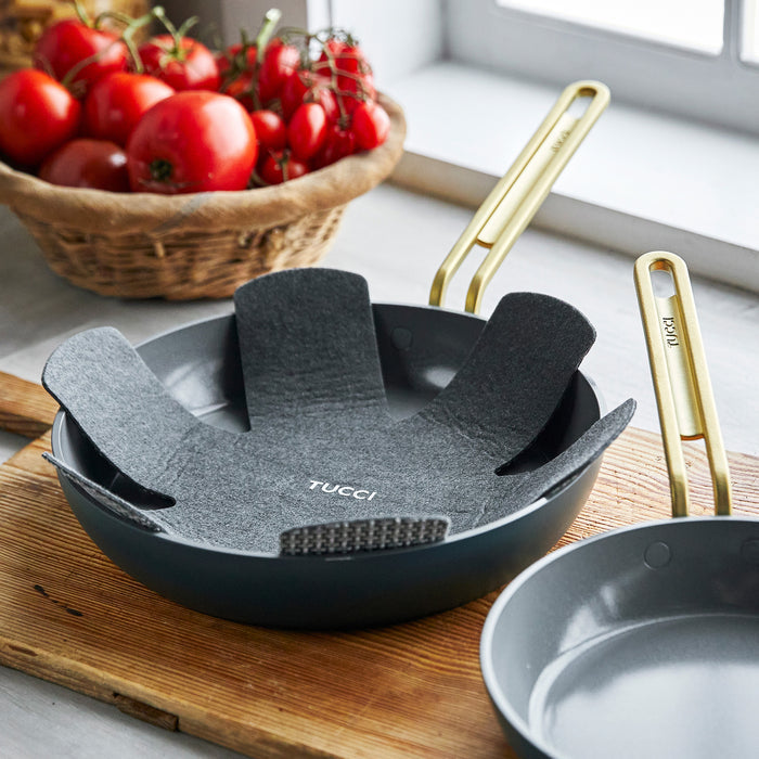 A Stanley Tucci™ Ceramic Nonstick 8" and 10" Frypan Set in Venetian Teal, with gold handles and protective felt liner, rests on wood near a window with fresh tomatoes, highlighting the elegance of Tucci cookware.