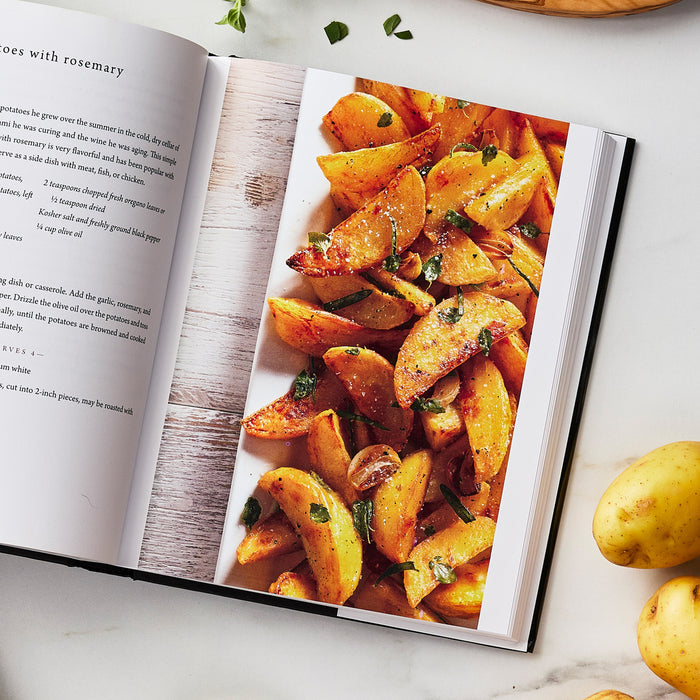 The Tucci Cookbook displayed open, showing a recipe page with golden roasted potato wedges topped with herbs.