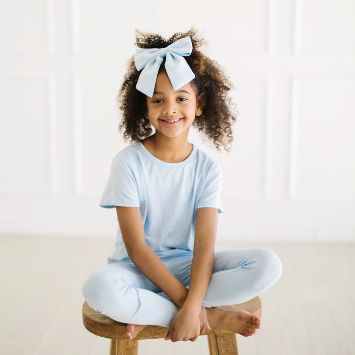 Young girl sitting on a wooden stool wearing the Toddler Basic Tee in Breeze and matching leggings with a blue bow in her hair