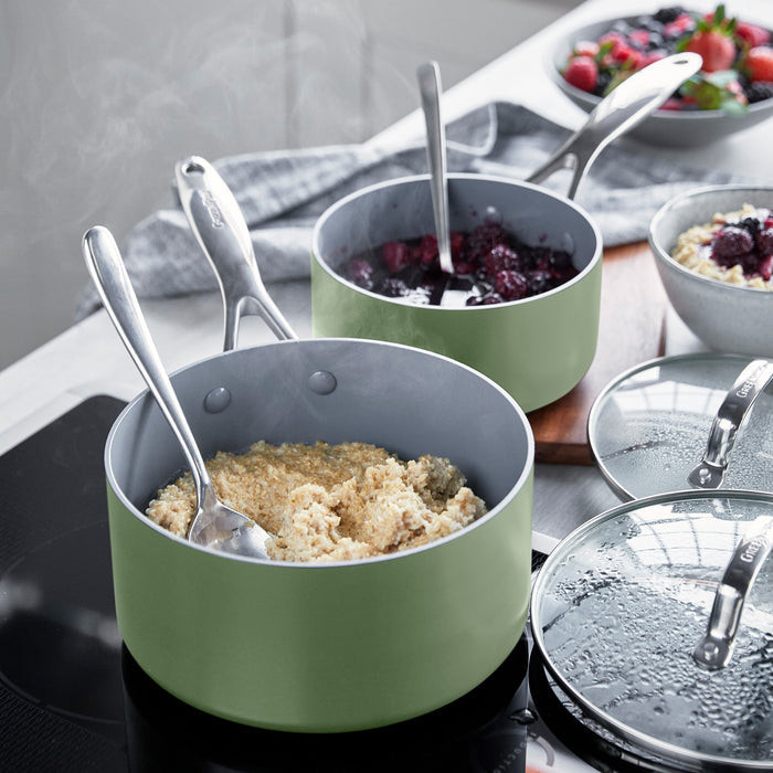 Two sage green ceramic saucepans with lids on a stove, one filled with oatmeal topped with berries. Set of two sizes.