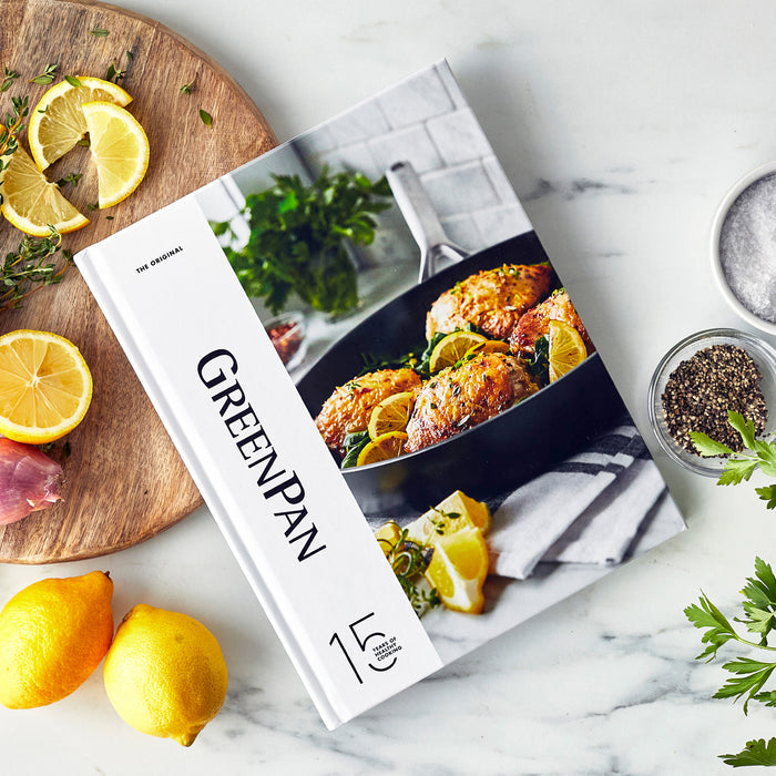 Hardcover 15-Year Anniversary Cookbook on marble with lemons, fresh herbs, salt, and pepper nearby.