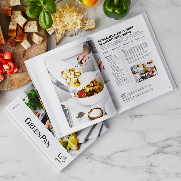 Cookbook open to a panzanella salad recipe with bread and fresh ingredients on a marble countertop.