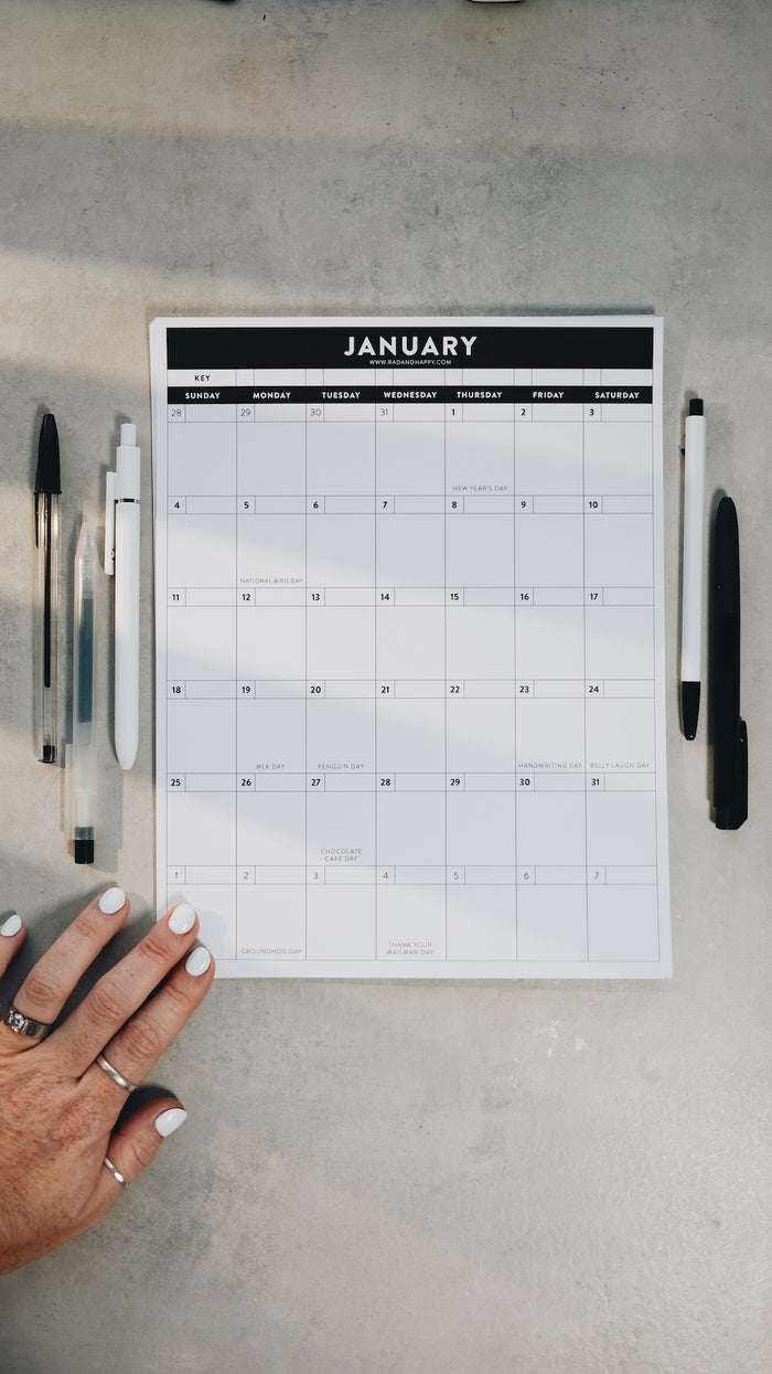 Hand holding a January calendar printout on a gray surface with pens.