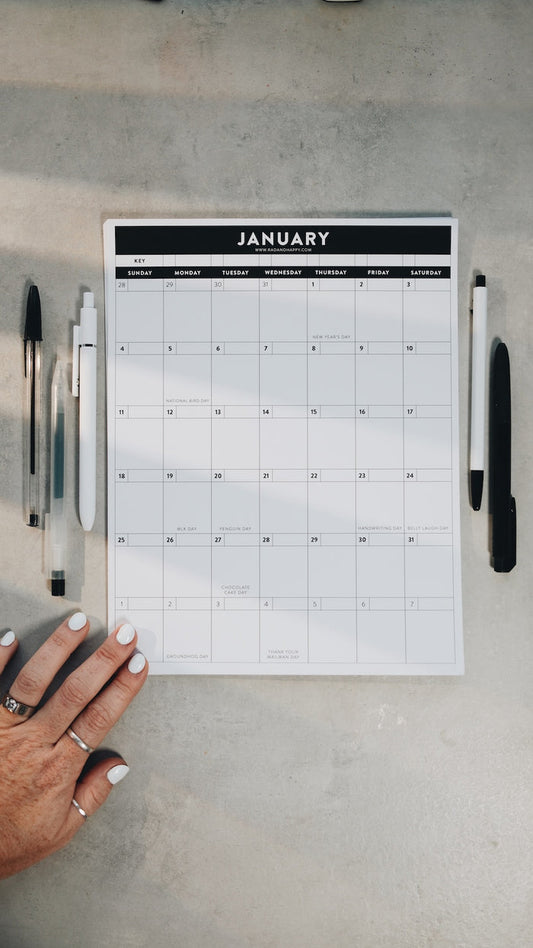 Hand holding a January calendar printout on a gray surface with pens.