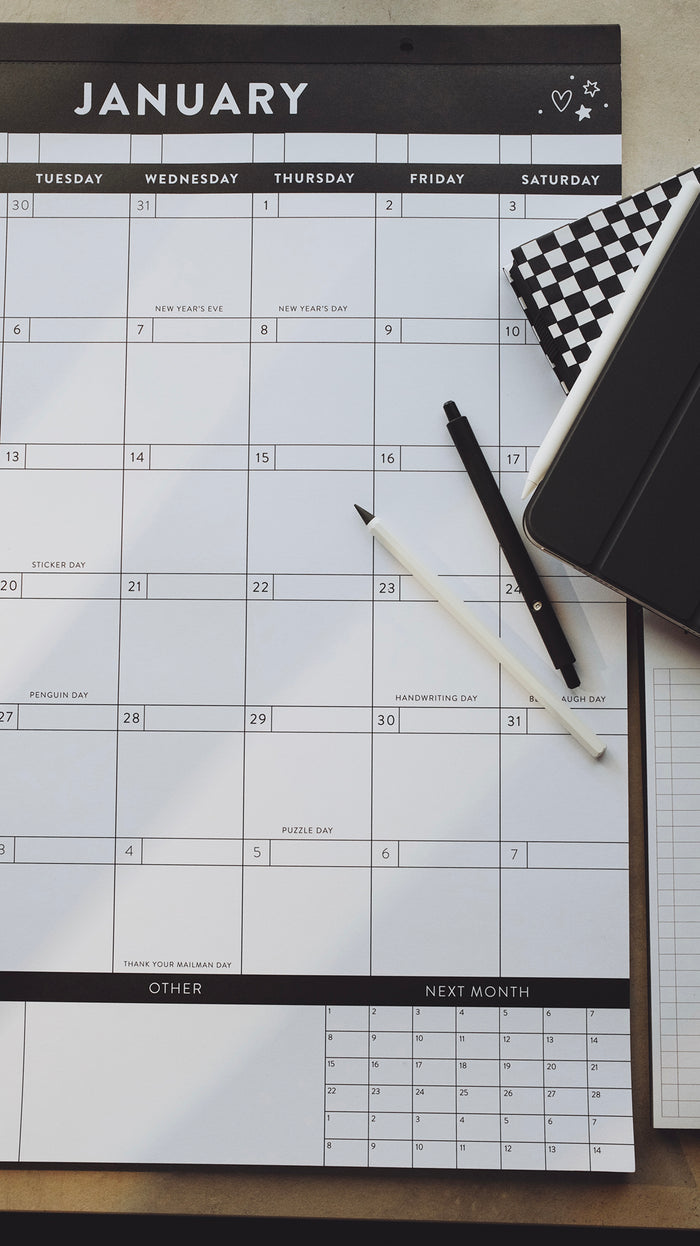 Pens and planners laying on top of a calendar showing the month of January on a light gray surface