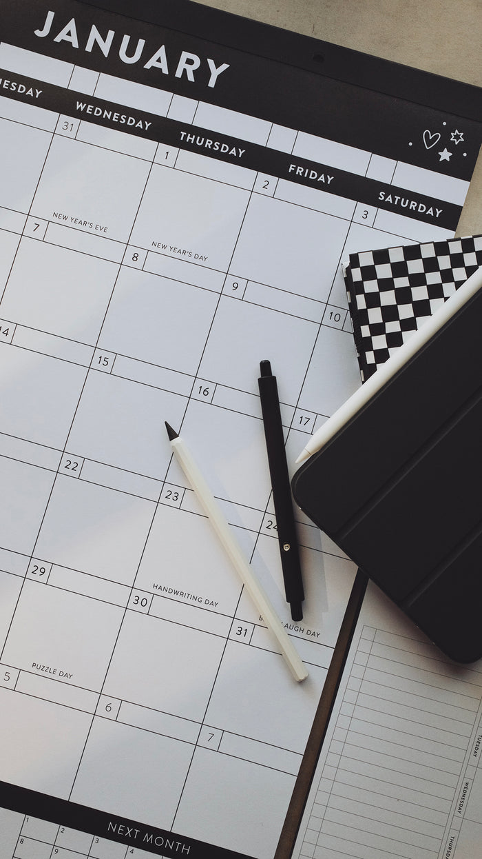 Pens and planners laying on top of a calendar showing the month of January on a light gray surface
