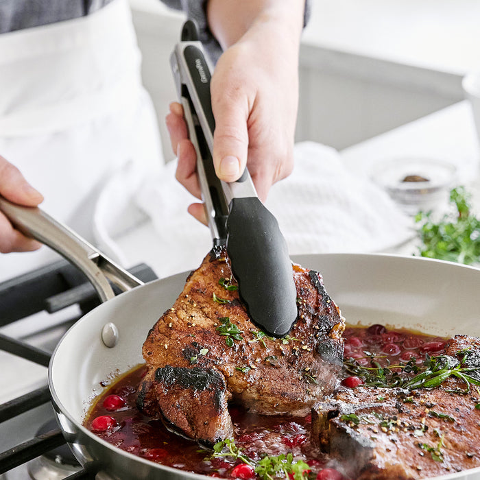 Black silicone utensil from a 5-piece set used to cook a pork chop with cranberries and herbs in a skillet.