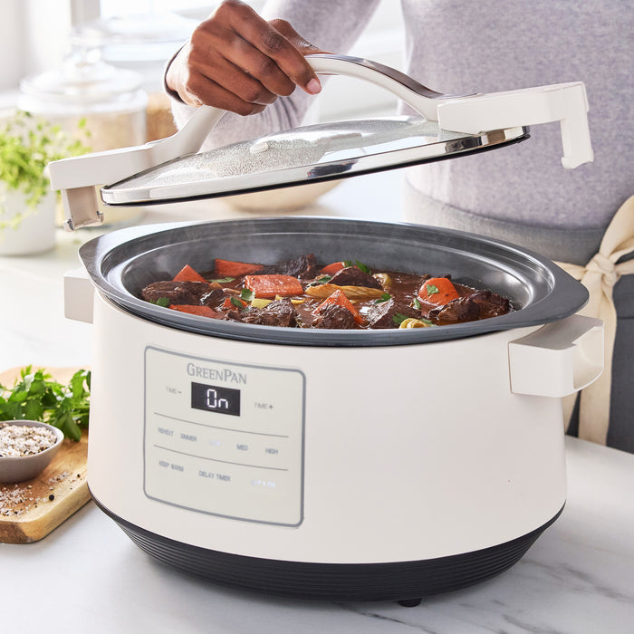 Person lifting lid of an 8-quart white slow cooker filled with stew, sitting on a kitchen counter.