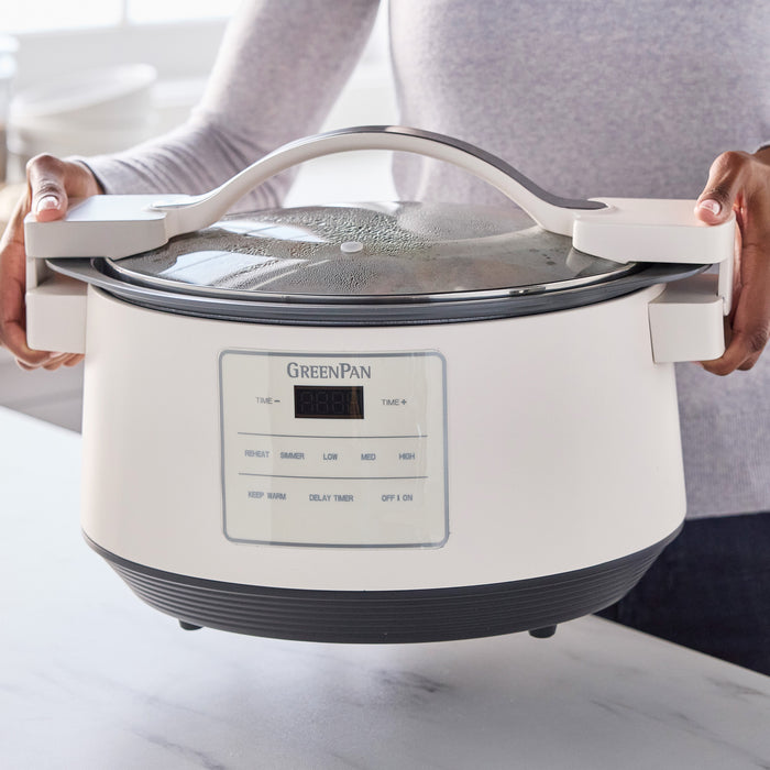 Person holding a cream-colored 8-quart slow cooker with a digital control panel on a kitchen counter.