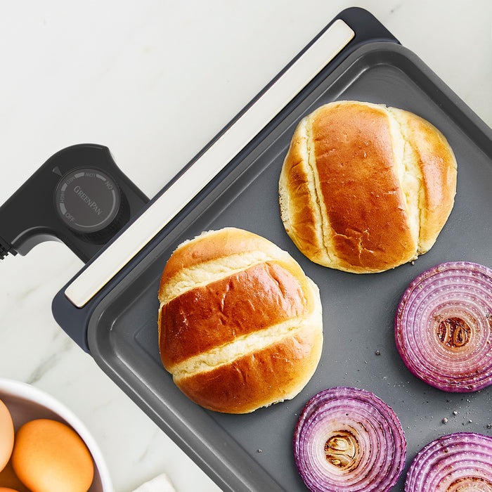 Two browned buns and red onion slices sizzle on the Advanced Griddle with Warming Drawer | Graphite, while a bowl of eggs sits nearby—a perfect setup for preparing a delicious family-sized brunch.