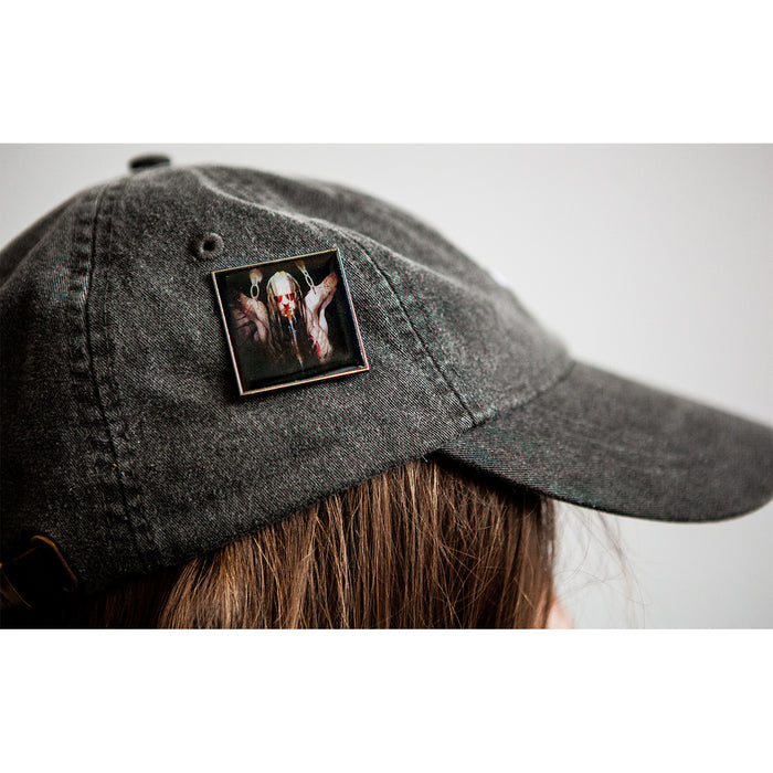 A person wears a dark gray baseball cap with a Sadistik enamel pin showing a long-haired, bearded man in dark clothing raising his hands near altars.