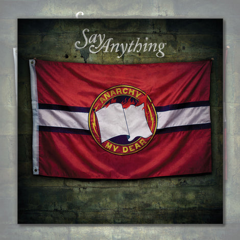 A red flag with a white horizontal stripe, blue borders, and a central emblem of an open book with flames and "Anarchy My Dear" hangs on the wall. Above it is “Say Anything,” matching the band's CD cover. Brand Name: Say Anything.