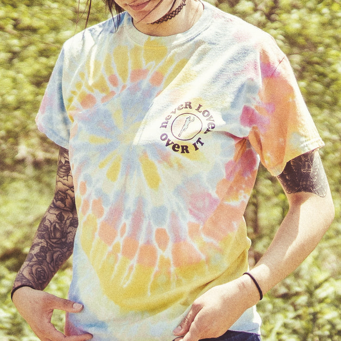 Multi-colored tie dye short sleeve with circular text surrounding a design in the top right corner. Text in the top right corner says NEVER LOVED, OVER IT. Model is wearing the shirt, standing in front of greenery.
