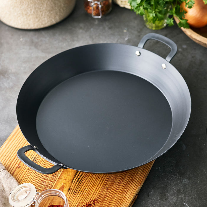 Carbon steel paella pan on a wooden board, surrounded by kitchen ingredients and fresh herbs.