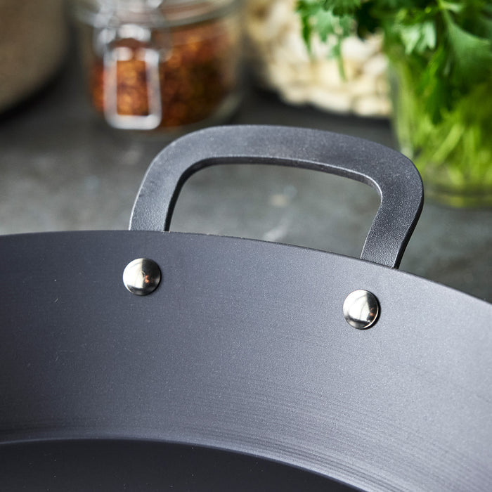 Carbon steel paella pan handle with glass jars and fresh green herbs in the blurred background.