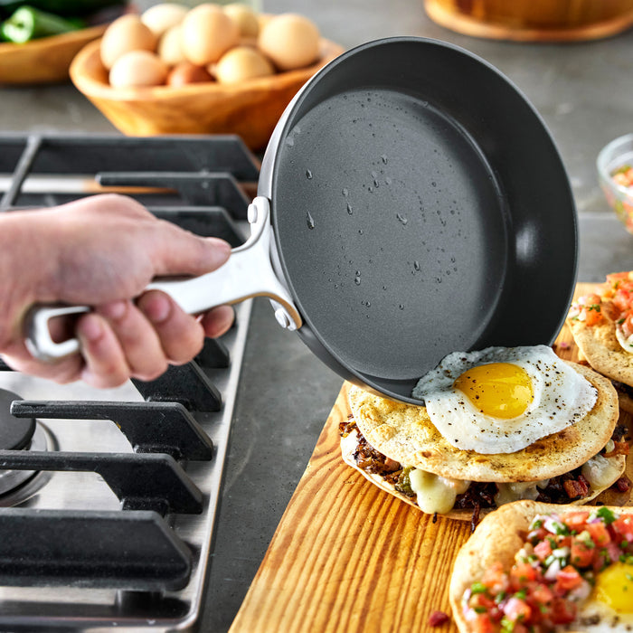 Hand slides fried egg from Bobby Flay by GreenPan Chef Series pan onto food near stove, with eggs on counter.