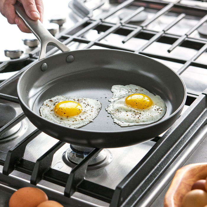 Two eggs frying in a gray hard anodized GreenPan skillet on a gas stove.