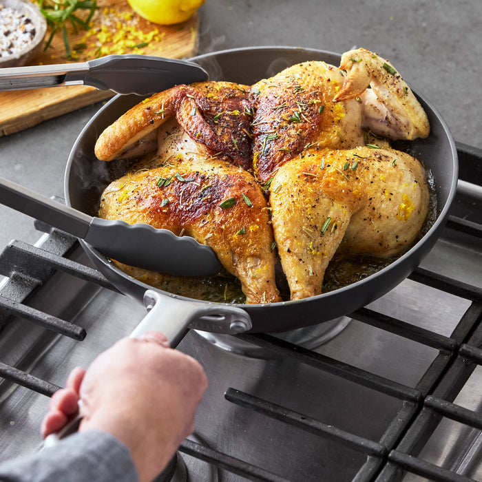 Spatchcocked chicken being cooked with tongs in a black Bobby Flay by GreenPan hard anodized skillet.