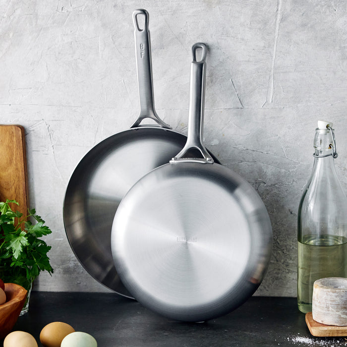 Two uncoated stainless steel frypans, 10-inch and 12-inch, on a gray kitchen counter.