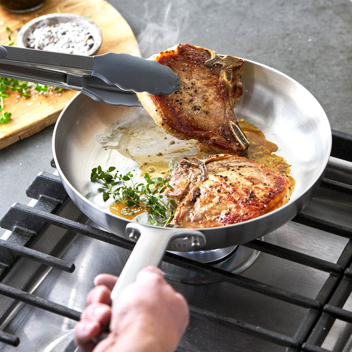Stainless steel frying pan with pork chops cooking on a stovetop.