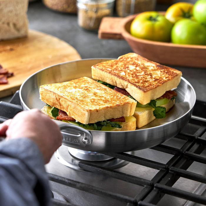 Two sandwiches in bacon, cheese, and greens cook in a Bobby Flay by GreenPan 10 and 12 frypan set on a stove.