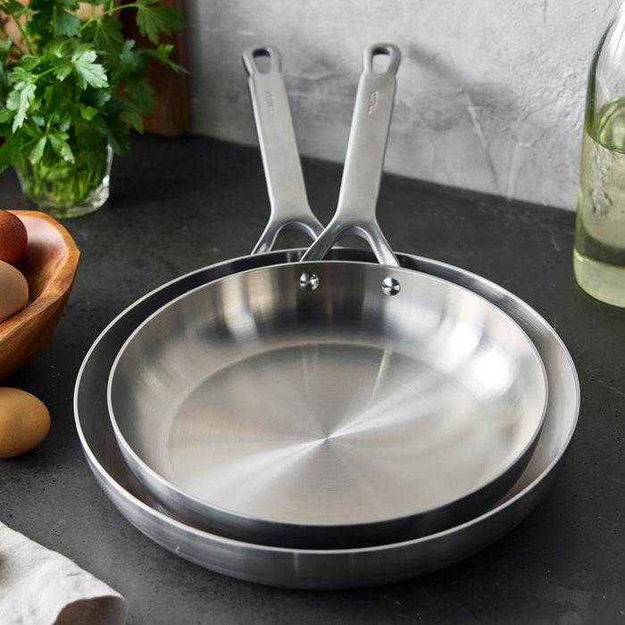 Two stainless steel frypans, 10 and 12, on a kitchen counter with eggs and parsley nearby.
