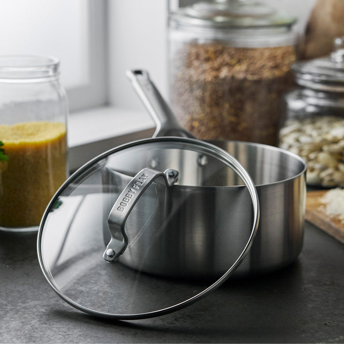 Stainless steel saucepan with glass lid on a counter near jars of grains.