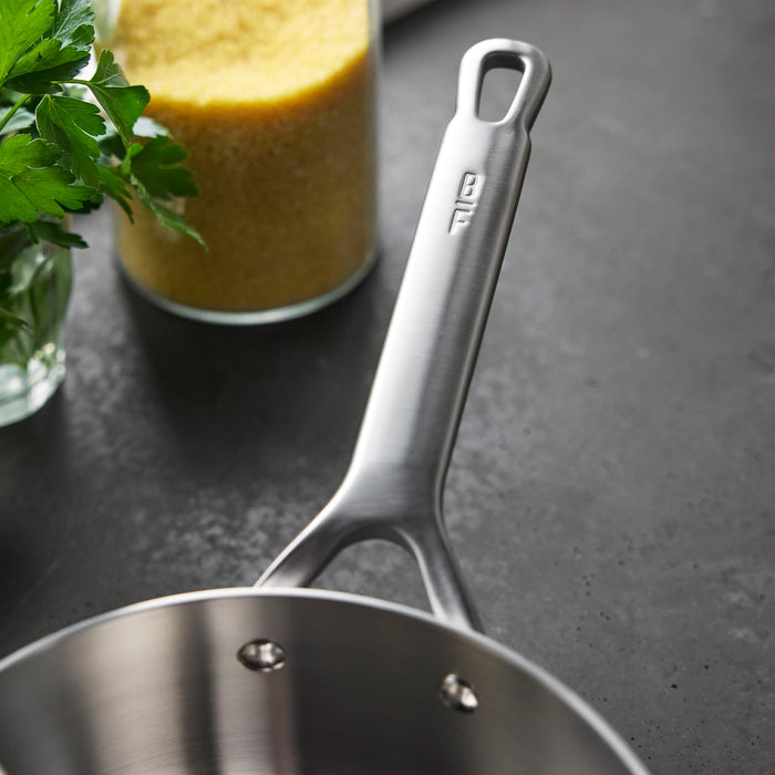 Stainless steel saucepan with lid on a dark counter, parsley and yellow grains beside it.