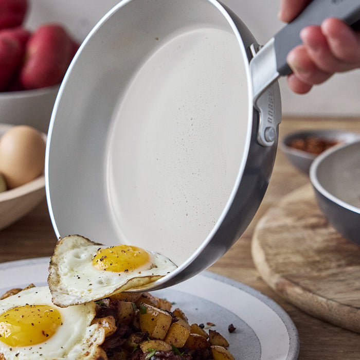 Fried egg sliding from a green ceramic nonstick frypan onto a plate with potatoes and another egg.