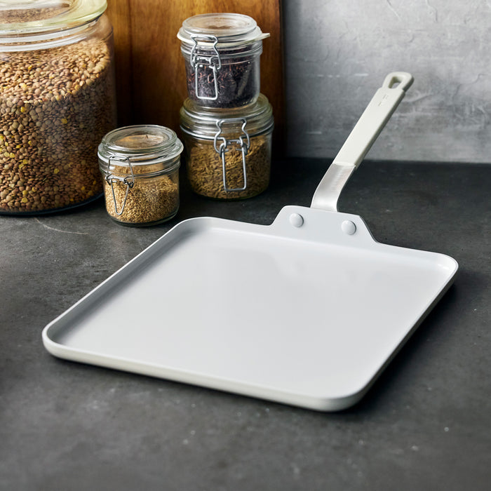 Square ceramic griddle with a handle on a countertop next to clear glass jars filled with dry ingredients.