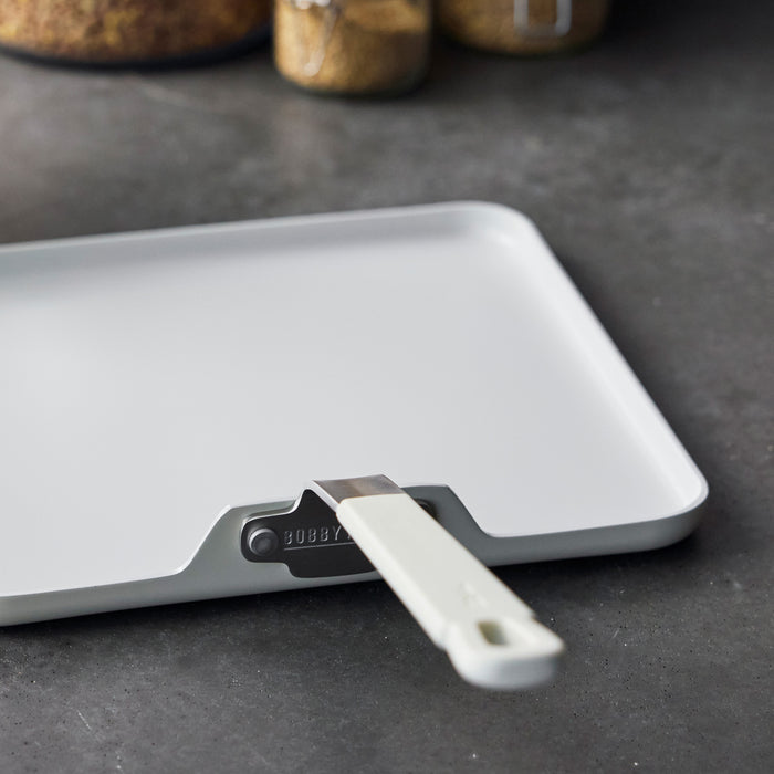 Cream ceramic square griddle with handle on a dark countertop, glass jars in the background.