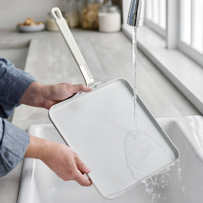 Cream ceramic nonstick square griddle being rinsed under running water at a kitchen sink.