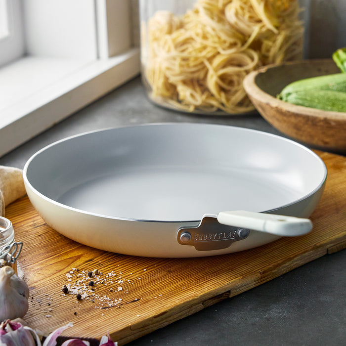 Cream-colored 12-inch frypan on a wooden board with pasta, zucchini, garlic, and spices nearby.