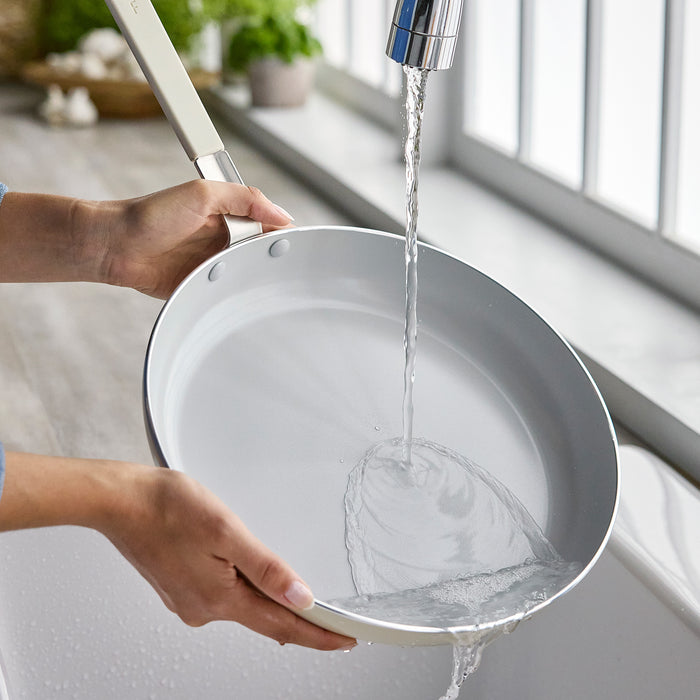 A hand rinses a black ceramic nonstick frypan in a bright kitchen sink.