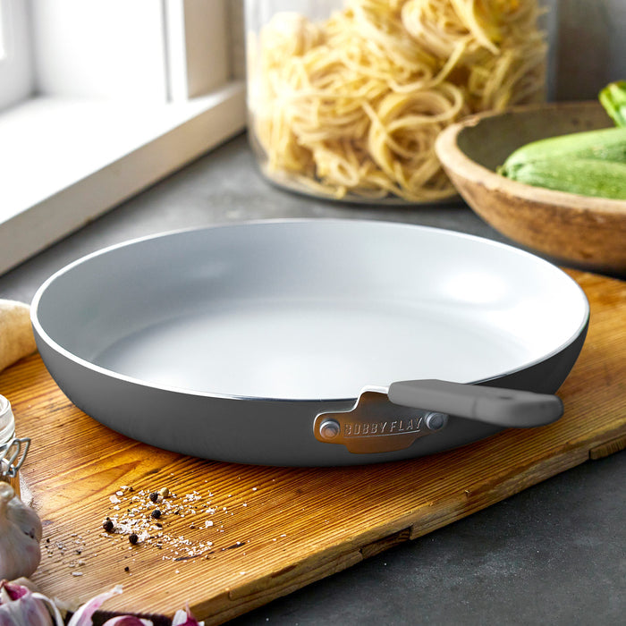 Ceramic nonstick frypan on a wooden board, with uncooked pasta, zucchini slices, and garlic cloves nearby.