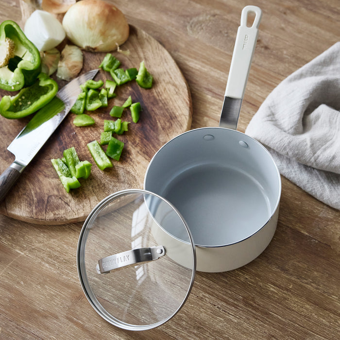 Cream ceramic saucepan with lid on a wooden board beside chopped vegetables.