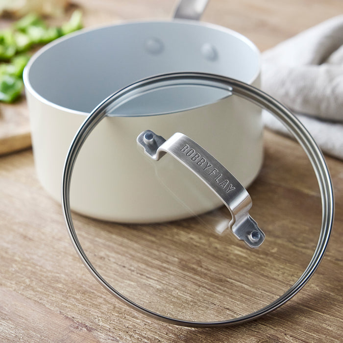 Cream ceramic 1.6-quart saucepan with lid, placed on wooden surface next to chopped greens.